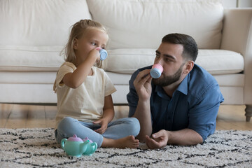 Caring dad and cute little daughter girl playing tea party, imagining drinking coffee, tea, using toy small plastic mugs, tiny expresso cups, enjoying game, playtime, family leisure on heating floor - Powered by Adobe