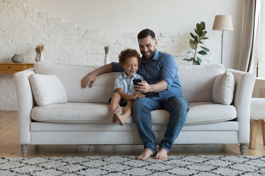 Happy dad hugging cheerful little son kid with love, care, holding smartphone, using cell for video call, taking selfie, sitting on home couch with boy. Father and child posing for picture