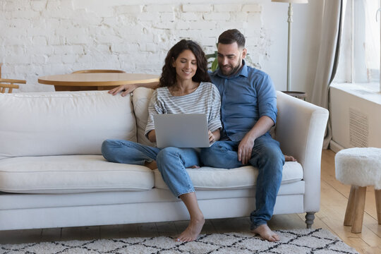 Happy engaged millennial couple in love sitting on couch at home, holding laptop on lap, sharing computer, using online shopping app, internet service, talking, chatting, smiling, hugging. Full length