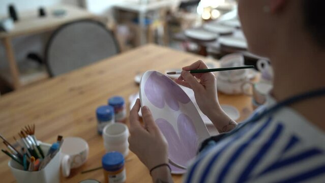 Woman painting and decorating ceramic object while visiting art class or workshop, ceramist holding paintbrush drawing color patterns on clay plate in her own pottery studio, over-the-shoulder shot 