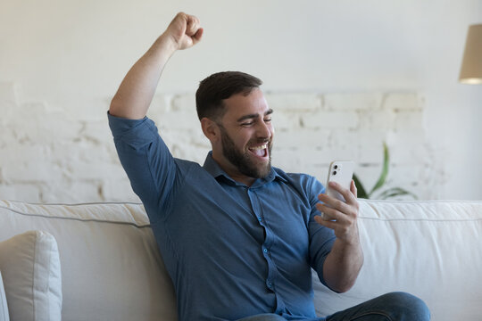 Excited happy smartphone user guy learning good news, reading text message, notice on screen, making winner yes hand with happy smile, laughing, shouting, sitting on home couch, celebrating achieve