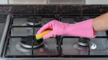 A woman's hand in a rubber glove washes a gas stove with a sponge