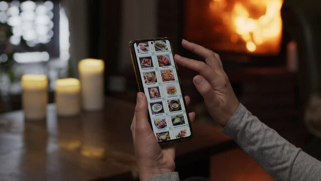 Ordering food using a smartphone at home. A woman selects restaurant food in the internet menu of a gourmet restaurant using an application on a smartphone. Home evening furnishings with a burning 