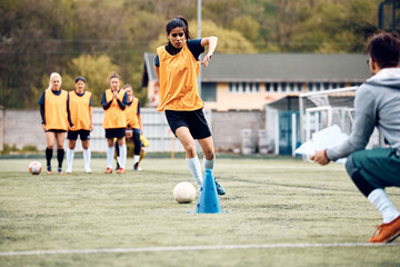 Female soccer player leading ball among cones during sports training.