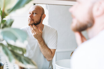 Handsome bald man looking at mirror and touching face in bathroom