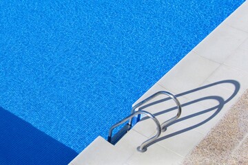 Detalle parcial de una piscina de agua transparente con la escalera de acceso y su sombra en primer plano.