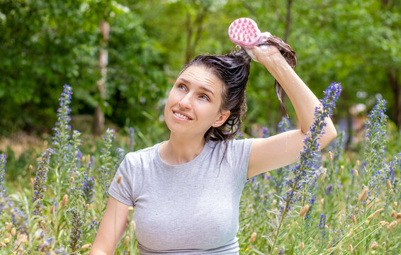 hair growth stimulating,scalp massage.woman in garden holding hair up with both hands,in pony tail,green grass,flowers background.shapoo foam on hair.pink scalp massager brush with silicone,flexible 