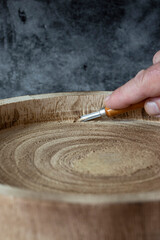 Man carving a wood tree log into a bowl or platter, with a beveled chisel.. On a dark stone...