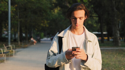 Young man in the park is typing a message. Guy walks in the park and typing a text message on a mobile phone. Male person looks into a cell phone and types a text sms. People and technology.