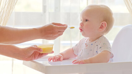 Mom feeds a 9 month baby with fruit puree from a spoon, close-up, high key.  Mother is Feeding a baby from a spoon.  Infant boy eats sitting on baby's chair. Mother cares about little son.