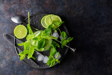 Mojito cocktail, lime slices, mint and ice on a dark background. Cocktail mojito, ice tongs and cocktail spoon on a black plate. Top view. Copy space