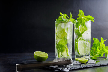Mojito cocktail, mint, lime slices and ice cubes on a dark background. Cocktail mojito, ice tongs and cocktail spoon on slate board. Copy space