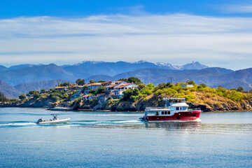 Tugboat drags a small motor boat on a rope against the backdrop of houses of a small town, mountains and blue sky