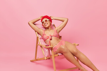 Happy relaxed young woman dressed in swimsuit bathhat and sunglasses poses on comfortable deck chair takes nap gets suntan enjoys summer vacation isolated over pink background. Perfect holiday - obrazy, fototapety, plakaty