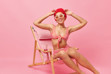 Happy woman wears bathhat sunglasses and striped bathing suit poses on comfortable deck chair enjoys summer vacation smiles broadly isolated over pink background. Relaxation and holiday concept