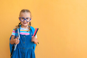 Little school girl in uniform and eyeglasses with backpack standing over yellow background, holding notebook and colored pencils, smiling