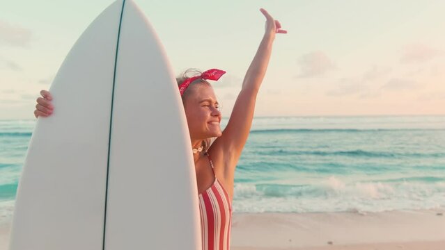 Happy young blonde Caucasian woman sends greetings to friends surfers showing shaka gesture inviting to joint vacation holds white surfboard in front stands on beach. Sports, lifestyle, travel