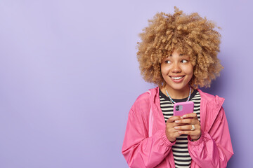 Positive curly haired young woman enjoys messaging on mobile phone dressed in casual clothes smiles happily focused away isolated over purple background with copy space for your advertisement