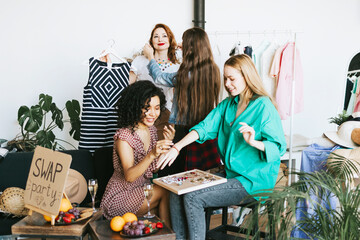 four young woman female caucasian and african students at swap party try on clothes, bags, shoes and accessories, change clothes with each other, second hand for things, zero waste life, eco-friendly