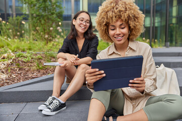 Happy mixed race female friends meet together take selfie on modern tablet prepare for exams dressed in casual clothes sit on stairs outdoors in city browse touchpad watch online movie content