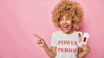 Horizontal shot of surprised cheerful young woman with curly hair holds two sanitary napkins with...