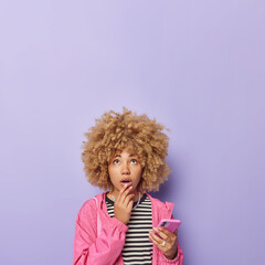 Indoor shot of surprised curly haired woman holds chin looks with shocked expression above holds cellular communicates online wears pink jacket striped jumper isolated over purple background