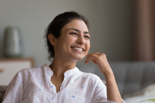 Happy beautiful young Indian woman looking at window with toothy smile, thinking of future vision, good news, dreaming, touching chin, facial skin, laughing. Head shot portrait