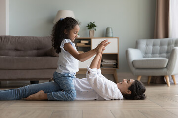 Happy cute little Indian kid girl sitting on mom on heating floor, clapping hands, giving high five. Young mother and child playing active games at home, doing exercises. Motherhood concept