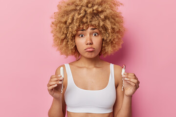 Horizontal shot of pretty displeased young woman with curly hair holds two tampons chooses best...