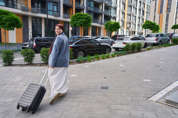 Woman stands in a half turn holding a suitcase on wheels