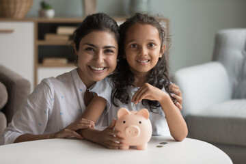 Happy beautiful Indian mother and kid girl saving money, holding piggy bank, looking at camera, smiling, laughing. Mom and child making reserve fund, investment, donations. Home portrait