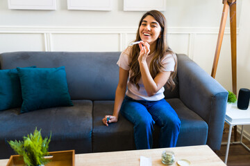 Excited young woman smoking weed and laughing in the living room - obrazy, fototapety, plakaty