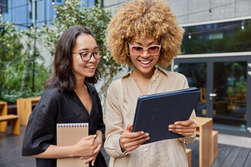 Positive mixed race friendly women focused at tablet carry spiral notebook stroll outdoors wait for feedback on their streaming video dressed in casual clothes stand outdoor in urban setting.