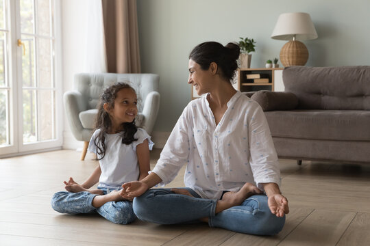 Happy Indian mom and kid girl enjoying yoga at home, sitting in lotus pose on heating floor, keeping zen fingers, talking, laughing, smiling. Mother teaching little daughter to meditate at home