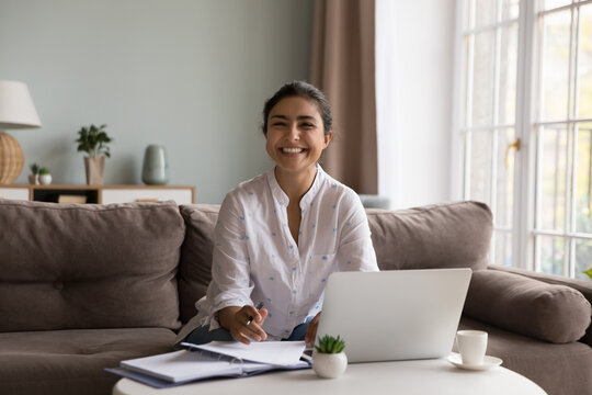 Happy young Indian woman working at laptop from home, writing notes, checking paper documents, doing paperwork, looking at camera, smiling, laughing, sitting on couch at computer. Home portrait