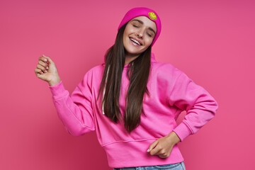 Obraz na płótnie Canvas Beautiful young woman in hooded shirt and funky hat dancing against pink background