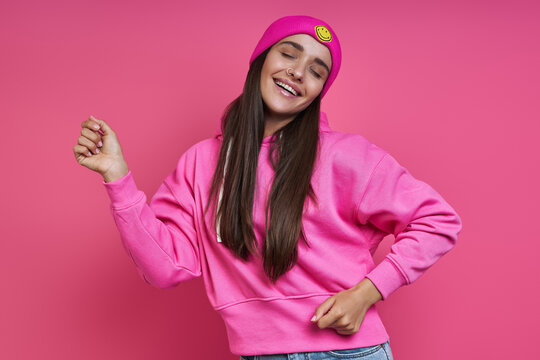 Beautiful young woman in hooded shirt and funky hat dancing against pink background
