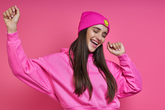 Happy young woman in hooded shirt and funky hat dancing against pink background