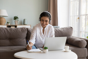 Happy millennial Indian student woman in wireless headphones writing learning notes, doing training course task, college homework, watching webinar, listening tutorials on laptop - obrazy, fototapety, plakaty