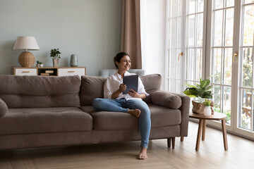 Happy Indian gadget user woman holding digital pad, resting on couch, looking at window away in...