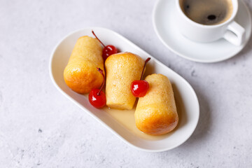 Neapolitan Rum baba (or baba au rum) on a white plate with a cocktail cherry and a cup of coffee. Small yeast cakes soaked in rum syrup on a gray background. Traditional Italian pastry. Close-up.