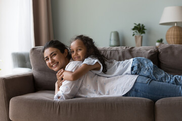 Happy sweet Indian kid girl resting on happy moms back, smiling. Cheerful pretty young mother lying...