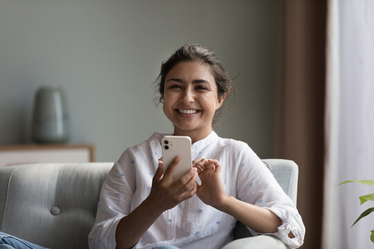 Happy beautiful millennial Indian woman using smartphone, looking at camera, smiling. Head shot portrait. Young cellphone user chatting, shopping on Internet, using online app