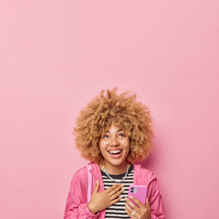 Vertical shot of positive woman with curly bushy hair keeps hand on chest smiles gladfully dressed in striped jumper and jacket holds mobile phone hears something funny isolated over pink wall