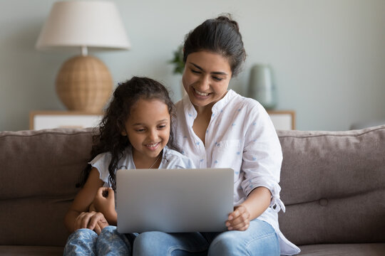 Happy pretty Indian mother and cheerful cute girl kid sharing laptop, enjoying leisure on couch together, watching movie, talking on video call, looking at monitor, smiling, laughing, hugging