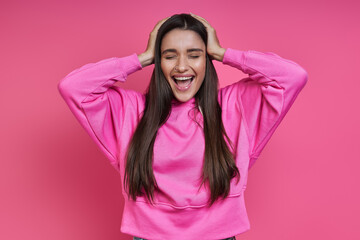 Happy young woman holding head in hands and keeping eyes closed against pink background