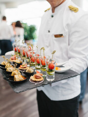 The waiter holds the appetizers on a black slate stand—pancakes with red caviar, Caprese in a glass on skewers.