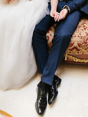 The bride and groom are sitting on a red sofa with a gold pattern holding hands. Legs of a man in a blue suit close-up.