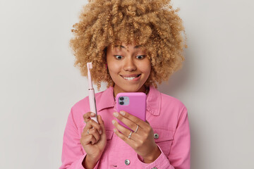 Beautiful woman with blonde curly hair bites lips focused into smartphone screen searches information how to care about teeth holds electric toothbrush dressed in pink jacket isolated on grey wall