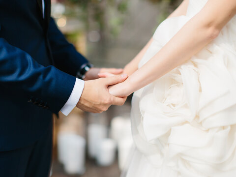 Newlyweds hold hands close-up. The groom is in a dark blue suit, the bride in a white airy dress with flowers.